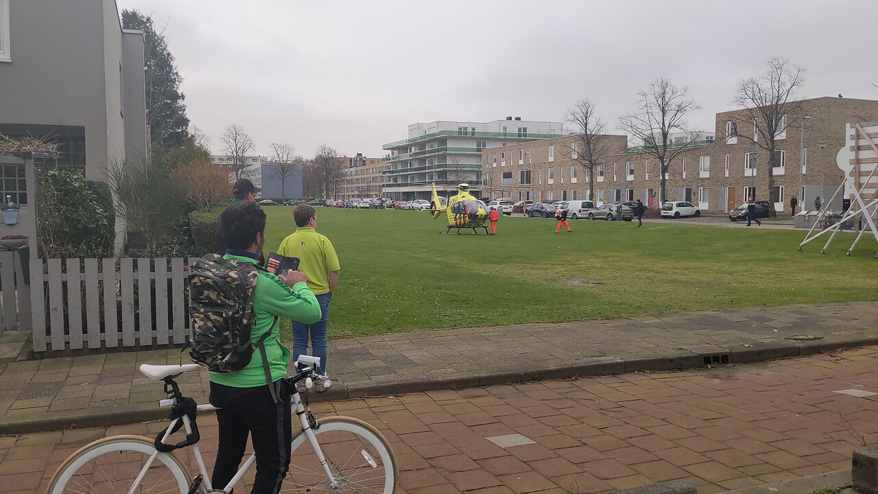 Ambulance helicopter landed on a grass field in a residential area, bystanders on the pavement away from the landing area