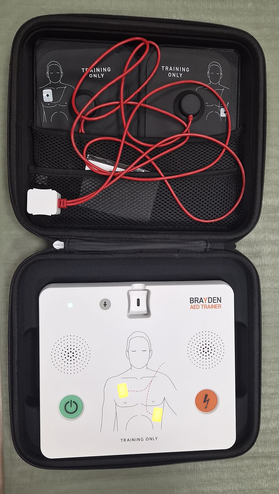 Training AED in a protective case with training pads, showing pad diagrams and the shock button